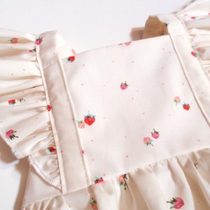 May include: Close-up of a cream-colored fabric with a strawberry pattern. The fabric features small, red strawberries with green leaves and pink dots. The design includes ruffled edges and straps.