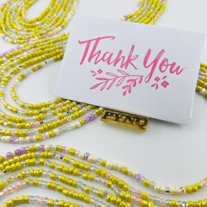May include: A multi-strand waist bead necklace with yellow, white, pink, and silver beads. The necklace is laid out on a white surface with a small card that says "Thank You" in pink lettering. The card also has a small gold charm with the letters "PYNO" on it.