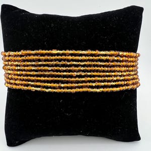 May include: A stack of amber-colored beaded bracelets with gold accents. The bracelets are arranged in a circle on a black velvet cushion.