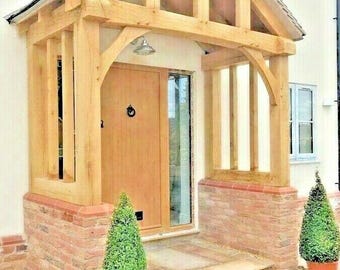 May include: A wooden porch with a gabled roof over a front door. The porch is supported by wooden beams and has a brick base. Two small evergreen trees are planted in pots on either side of the door.