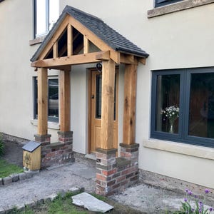 May include: A wooden porch with a gable roof over the front door. The porch has brick pillars and a wooden frame. The door is a solid wood door with a glass panel. The house is painted white with black windows.