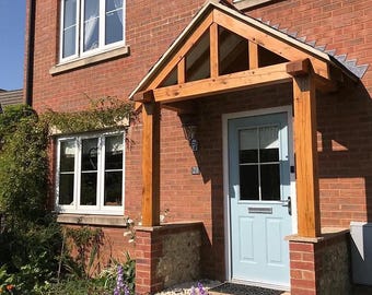 May include: A two-story brick house with a light blue front door and a wooden porch. The house has white-framed windows and a garden with colorful flowers. The porch is supported by wooden beams and brick pillars.