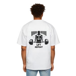 Op de afbeelding: Wit T-shirt met een zwarte afbeelding van een gewichtheffer die een halterstang optilt. De afbeelding bevat de tekst "Lift Repeat".