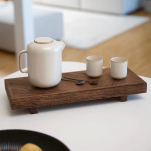 Può includere: Una teiera in ceramica bianca con coperchio e manico è appoggiata su un vassoio di legno scuro. Due piccole tazze bianche con bordi dorati e due cucchiaini sono anche sul vassoio. Il vassoio è su un tavolo bianco.