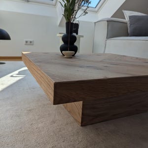 May include: A rectangular, light brown wooden coffee table with a unique, stepped design. A black vase with green and brown plants sits on the table. The table is in a well-lit room with a light-colored rug.