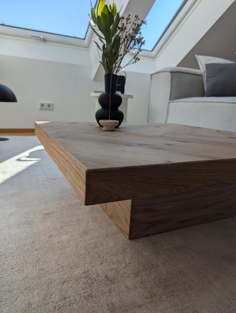 May include: A rectangular, light brown wooden coffee table with a unique, stepped design. A black vase with green and brown plants sits on the table. The table is in a well-lit room with a light-colored rug.