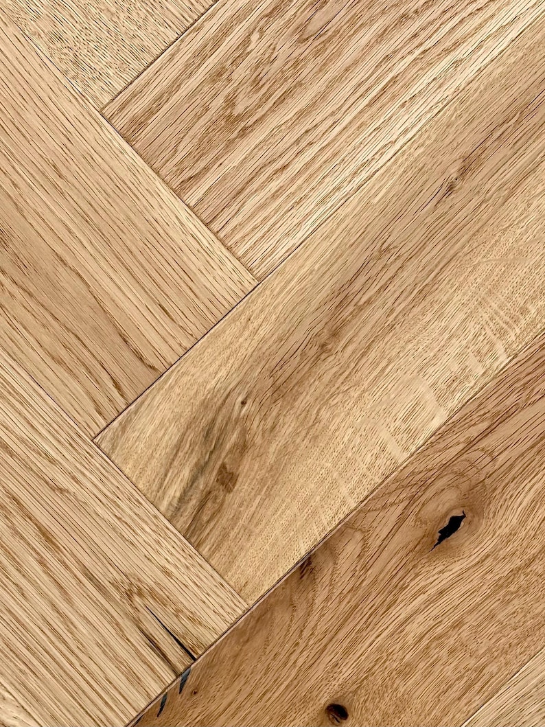 May include: Close-up of a herringbone pattern wood floor. The wood planks are a light brown color with visible grain and knots. The planks are arranged in a diagonal, zig-zag pattern, creating a textured surface. This flooring is suitable for home decor.