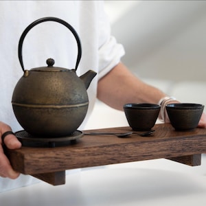 Può includere: Una teiera in ghisa color bronzo con manico nero e due tazze abbinate poggiano su un vassoio di legno. La teiera è rotonda con un piccolo beccuccio. Le tazze sono piccole e scure.