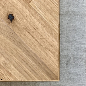 May include: Close-up of a wooden table with a herringbone pattern. The table is made of light-colored wood with visible grain and a knot. The table is set against a gray textured surface.
