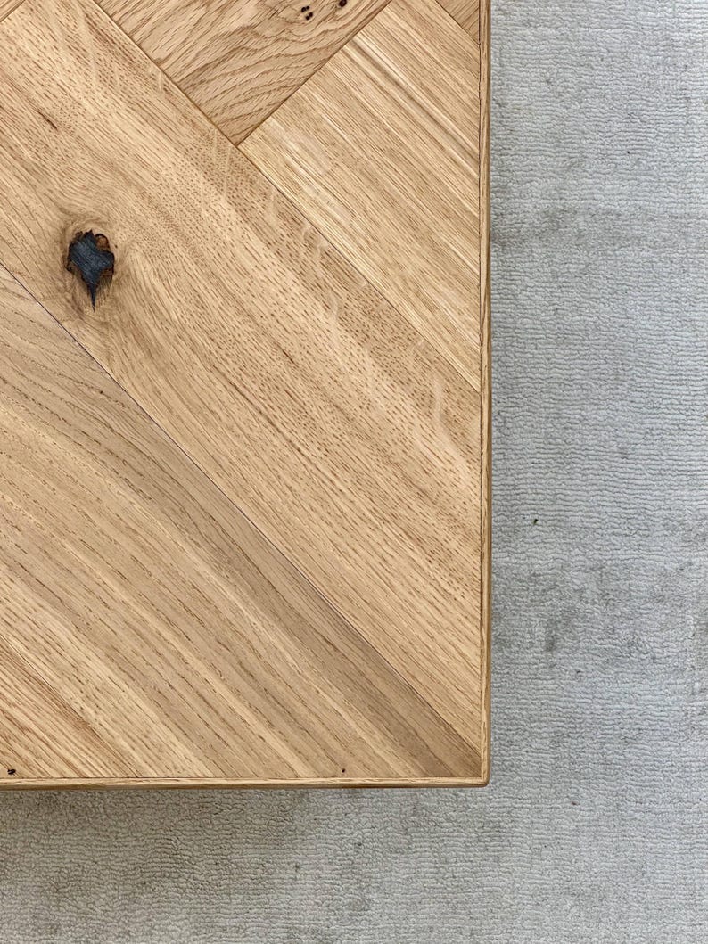 May include: Close-up of a wooden table with a herringbone pattern. The table is made of light-colored wood with visible grain and a knot. The table is set against a gray textured surface.