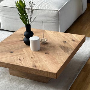 Peut inclure: Une table basse carrée en bois brun clair avec un fini grain de bois naturel. Un vase noir avec de la verdure, une bougie blanche et un porte-encens sont posés sur la table. La table est sur un tapis gris clair.