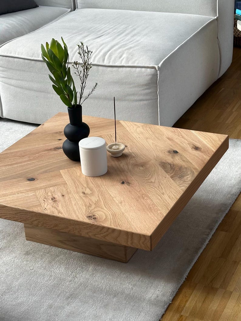 May include: A square, light-brown wooden coffee table with a natural wood grain finish. A black vase with greenery, a white candle, and an incense holder sit on the table. The table is on a light gray rug.