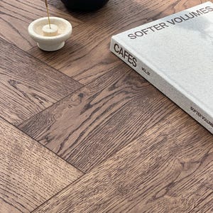 May include: A close-up of a wooden surface with a herringbone pattern. A book titled "SOFTER VOLUMES" and "CAFES VOL. 01" is on the surface. A black vase and a white incense holder with an incense stick are also present.