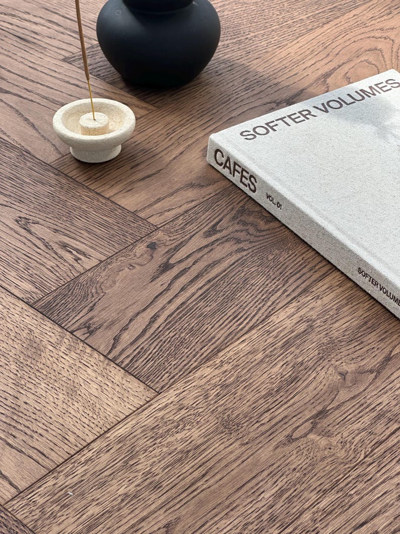 May include: A close-up of a wooden surface with a herringbone pattern. A book titled "SOFTER VOLUMES" and "CAFES VOL. 01" is on the surface. A black vase and a white incense holder with an incense stick are also present.
