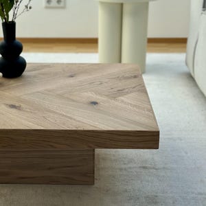 May include: A light brown wooden coffee table with a herringbone pattern on top. The table has a rectangular shape and a lower, rectangular base. A black vase with greenery sits on the table. A small, off-white side table is in the background.
