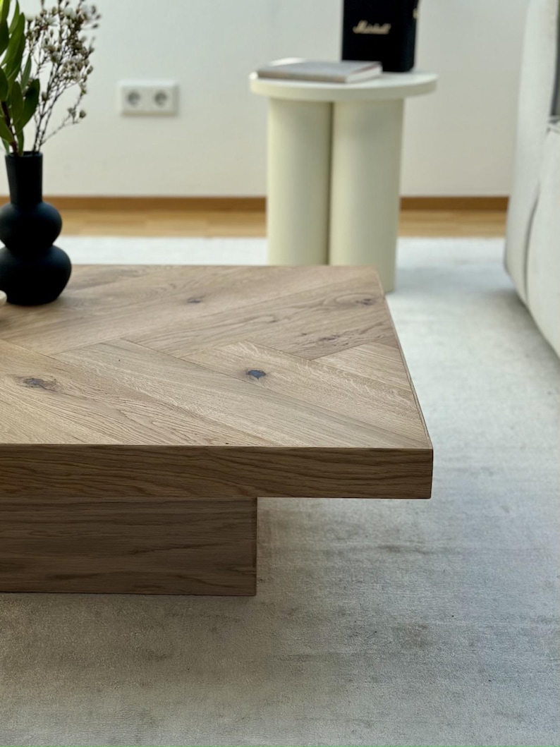 May include: A light brown wooden coffee table with a herringbone pattern on top. The table has a rectangular shape and a lower, rectangular base. A black vase with greenery sits on the table. A small, off-white side table is in the background.