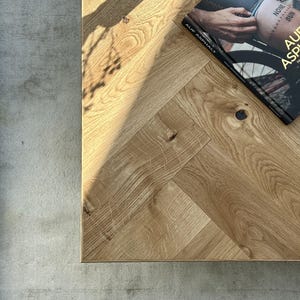 May include: A close-up shot of a light oak wood table with a herringbone pattern. A book titled "AUF ASPHALT" is on the table. The book cover features a person's leg with a tattoo. The table is next to a gray rug.