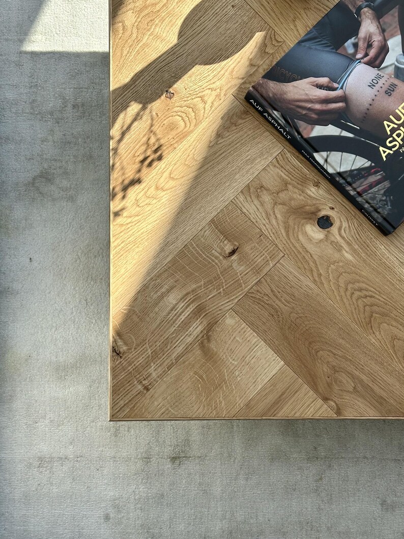 May include: A close-up shot of a light oak wood table with a herringbone pattern. A book titled "AUF ASPHALT" is on the table. The book cover features a person's leg with a tattoo. The table is next to a gray rug.