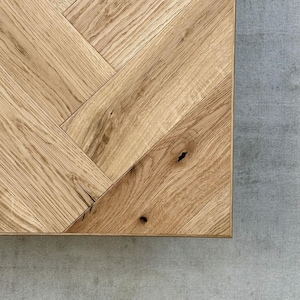 May include: Close-up of a corner of a light brown wooden table with a herringbone pattern. The wood grain is visible, and the table is set against a gray concrete surface.