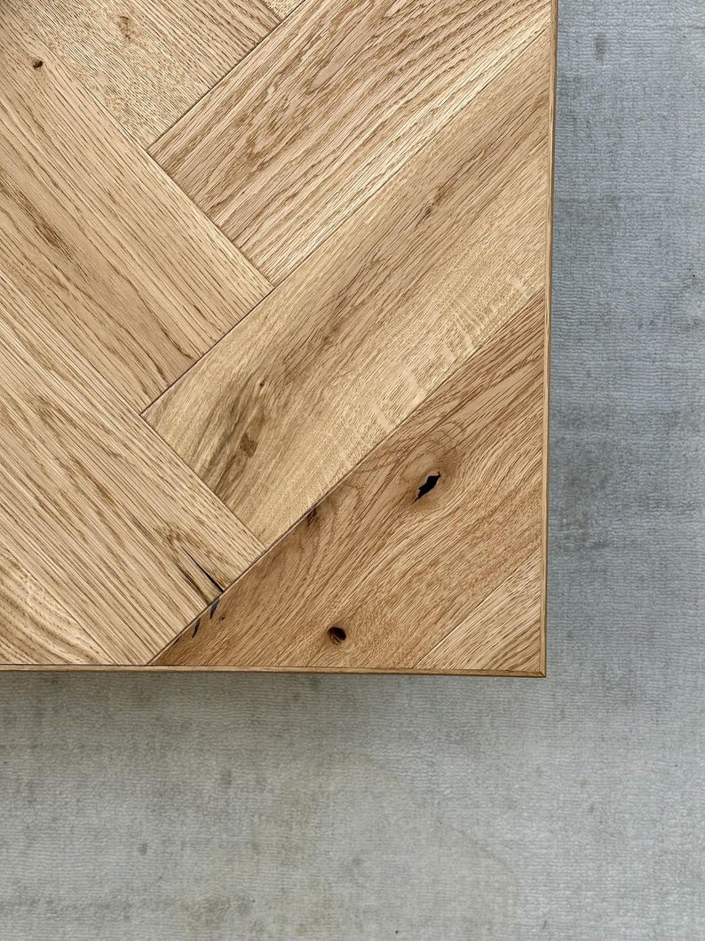 May include: Close-up of a corner of a light brown wooden table with a herringbone pattern. The wood grain is visible, and the table is set against a gray concrete surface.