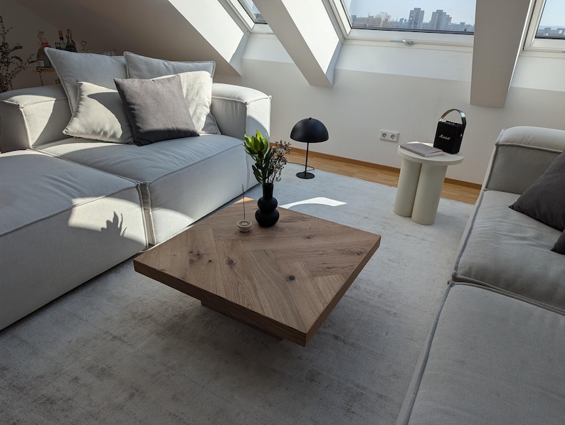 May include: A modern living room with a light gray sectional sofa, a wooden coffee table, and a small side table. The coffee table has a herringbone pattern. A black lamp and a small speaker are also visible.
