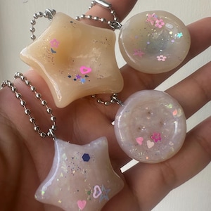 Puede incluir: Cuatro dijes de resina en cadenas de bolas plateadas. Dos dijes tienen forma de estrella en tonos melocotón translúcido y lavanda pálido, mientras que dos son circulares en blanco roto e iridiscente. Cada pieza contiene purpurina, corazones y flores.