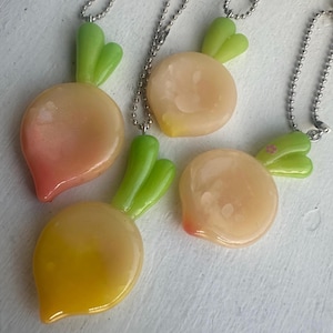 May include: Four charms shaped like round vegetables or fruits, each featuring a smooth, glossy finish in shades of peach, yellow, and translucent orange. Each charm has two bright green leaves at the top and hangs from a silver-toned ball chain.
