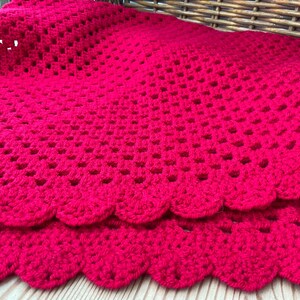 Puede incluir: Una manta de ganchillo de color rojo vibrante con un patrón clásico de cuadrados de la abuela y un diseño calado repetitivo. Los bordes del tejido están rematados con un borde festoneado decorativo, creando un aspecto suave y texturizado.