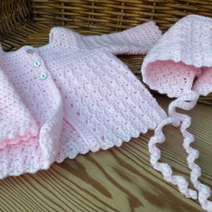 May include: A light pink, hand-crocheted baby set, including a cardigan with three buttons and a bonnet with ties. The set is made of soft yarn and features a delicate, openwork stitch. The bonnet has a scalloped edge.