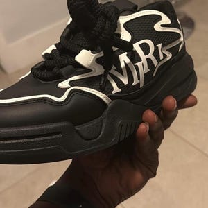 May include: Black and white sneaker with thick sole. The shoe features the letters "MR" in white on the side. The laces are black and thick. The shoe is being held by a person.