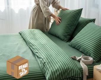 May include: Green striped bedding set with a duvet cover, fitted sheet, and pillowcases. The set is displayed on a wooden bed frame. The image includes a person arranging the pillows. The text "READY TO SHIP" is on a box.