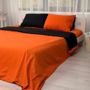 Puede incluir: Un juego de cama que incluye una sábana encimera de color naranja vibrante y una sábana bajera a juego, combinado con una funda nórdica negra en contraste. El juego incluye dos fundas de almohada rectangulares, una negra y otra naranja.