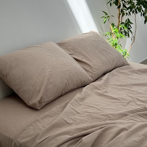 May include: A beige linen bed set, comprising two pillowcases and a fitted sheet. The bedding has a slightly creased texture. The bed is set against a white wall, with a houseplant visible in the background, creating a calming aesthetic.