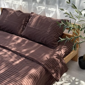 May include: A bedding set featuring a dark brown and light brown striped pattern. The set includes a duvet cover and two matching pillowcases, all displaying consistent horizontal stripes. The fabric is draped over a light-coloured wooden bed frame.