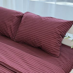 May include: A burgundy bed set with a striped design. The set comprises a fitted sheet and two pillowcases. The fabric has a slight sheen, and the stripes are horizontal across the pillowcases and the fitted sheet.