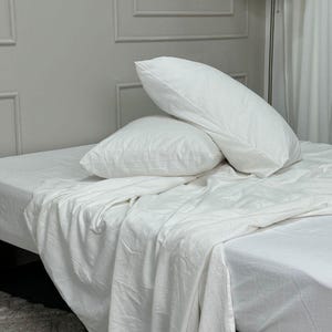 May include: A bed with white bedding, including a fitted sheet, a top sheet, and two pillows. The pillows are fluffy and rectangular. The sheets and pillowcases are made of a soft, textured fabric. The bed is in a room with a neutral color palette.
