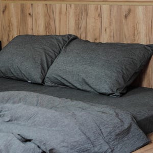 May include: A bed with gray bedding, including two pillows and a folded duvet. The bed is set against a wooden headboard and a wooden bed frame. The bedding appears to be made of a soft, textured fabric.