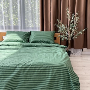 a bedroom with a bed covered in a green and white striped comforter and pillows. A small potted plant is placed next to the bed, and the room has brown curtains and a window with white curtains.
