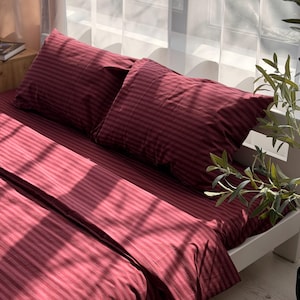 May include: A set of deep burgundy bedding featuring a subtle, repeating vertical stripe pattern. The set includes two rectangular pillowcases and a matching flat sheet draped over a bed frame, illuminated by natural light casting soft shadows.