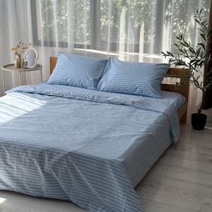 a neatly made bed with blue and white striped bedding, placed in a room with a window and a plant.