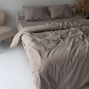 May include: A bed featuring a taupe and white pinstriped duvet cover and two matching rectangular pillowcases. The bedding is draped over a white wooden bed frame, creating a soft, layered appearance with visible fabric folds and creases.