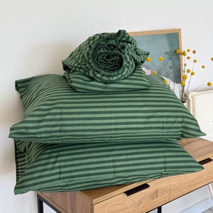 Puede incluir: Un juego de ropa de cama a rayas verdes, que incluye dos fundas de almohada y una sábana bajera enrollada. La ropa de cama es de color verde oscuro con rayas horizontales verdes más claras. Los artículos están apilados sobre una mesita de noche de madera con marco de metal negro.