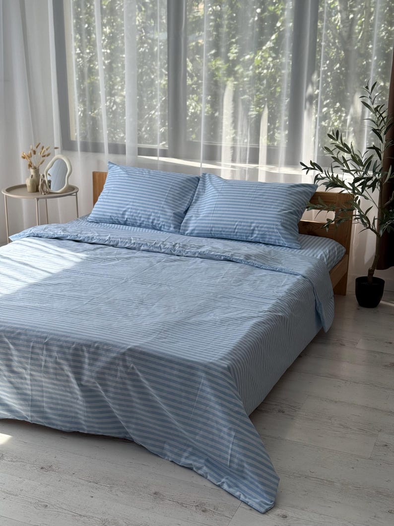 a neatly made bed with blue and white striped bedding, placed in a room with a window and a plant.