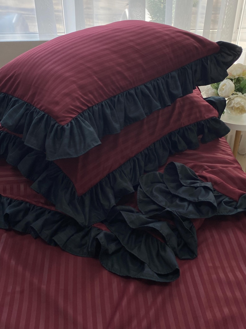 a bed with a red and black striped comforter and matching pillows, with a vase of white flowers on the windowsill.