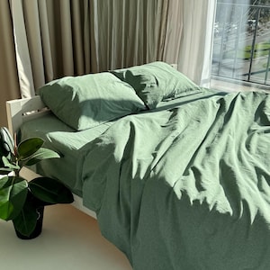 May include: A bed with sage green bedding, including a fitted sheet, two pillowcases, and a duvet cover. The bed is made of white wood and is positioned near a window with sheer curtains. A potted plant with large green leaves is in the foreground.