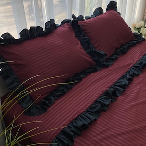 a bed with a burgundy-colored comforter and black ruffled trim, placed near a window with white curtains.