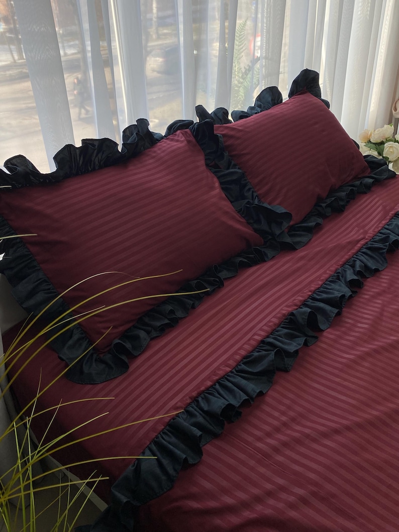 a bed with a burgundy-colored comforter and black ruffled trim, placed near a window with white curtains.