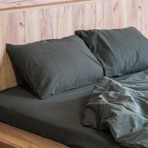 Könnte beinhalten: Dunkelgraue Bettwäsche, einschließlich zweier Kissen und eines Spannbetttuchs, auf einem Holzrahmen. Das Kopfteil besteht aus hellem Holz. Die Bettwäsche hat ein weiches, leicht zerknittertes Aussehen.