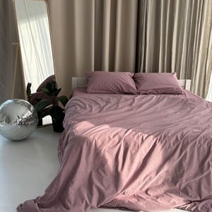 May include: A bed with a dusty rose duvet cover and two matching pillowcases. A full-length mirror, a disco ball, and a potted plant are in the background. The bed is against a neutral-toned curtain.