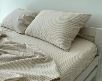 Può includere: Un set da letto composto da un lenzuolo con angoli e due cuscini rettangolari, realizzati in tessuto beige chiaro con sottili righe verticali marrone scuro. La biancheria da letto è disposta su una struttura per letto in legno bianco.